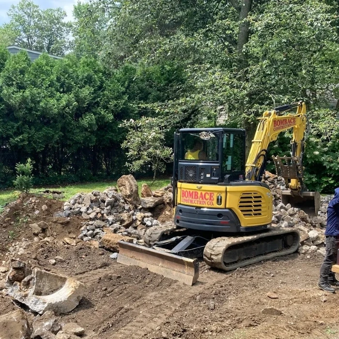 A Bombace Construction mini excavator clearing and grading a residential property site, surrounded by piles of broken concrete and rocks